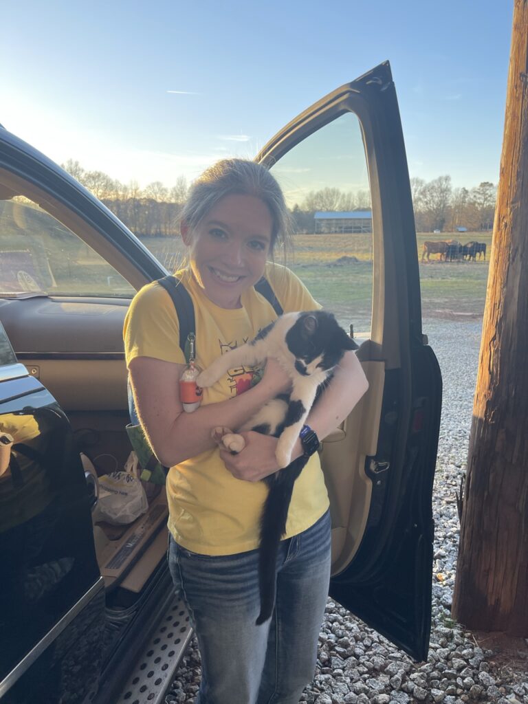 Woman smiling while holding a black and white cat, standing beside an open car door on a rural farm. She’s wearing a yellow shirt and jeans, with cows visible in the background under a clear blue sky.