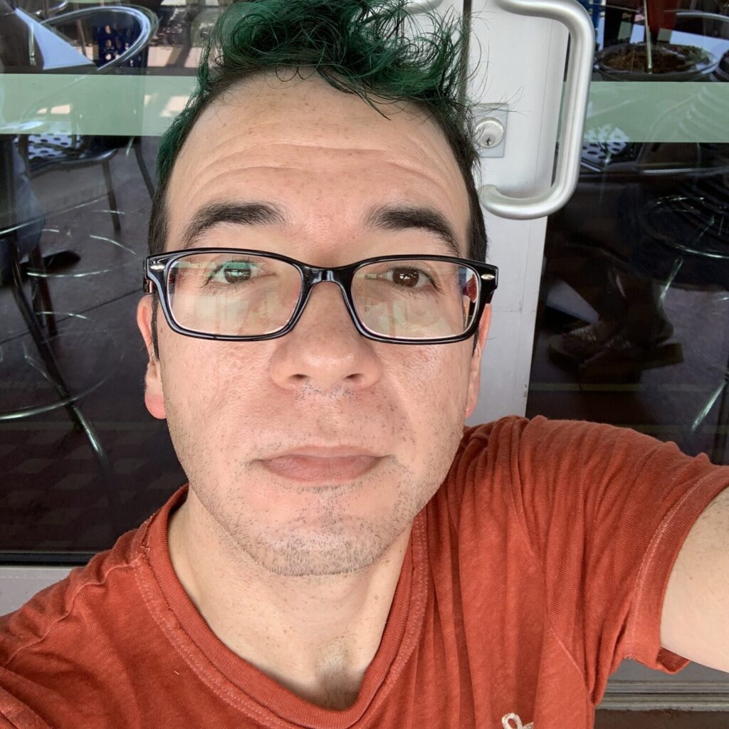 Selfie of a man with short green hair and black-rimmed glasses, wearing a red t-shirt, seated outside near a glass door with metal handles. Empty patio chairs and tables are visible in the background.